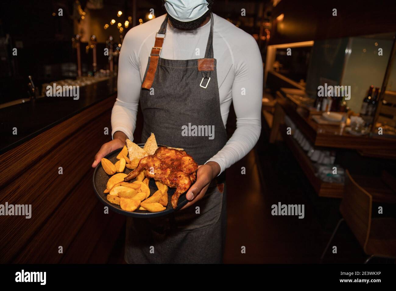 Afrikanischer Kellner mit einem Teller Huhn und Kartoffeln trägt Eine Coronavirus-Maske, um sich vor der Coronavirus-Pandemie zu schützen Stockfoto