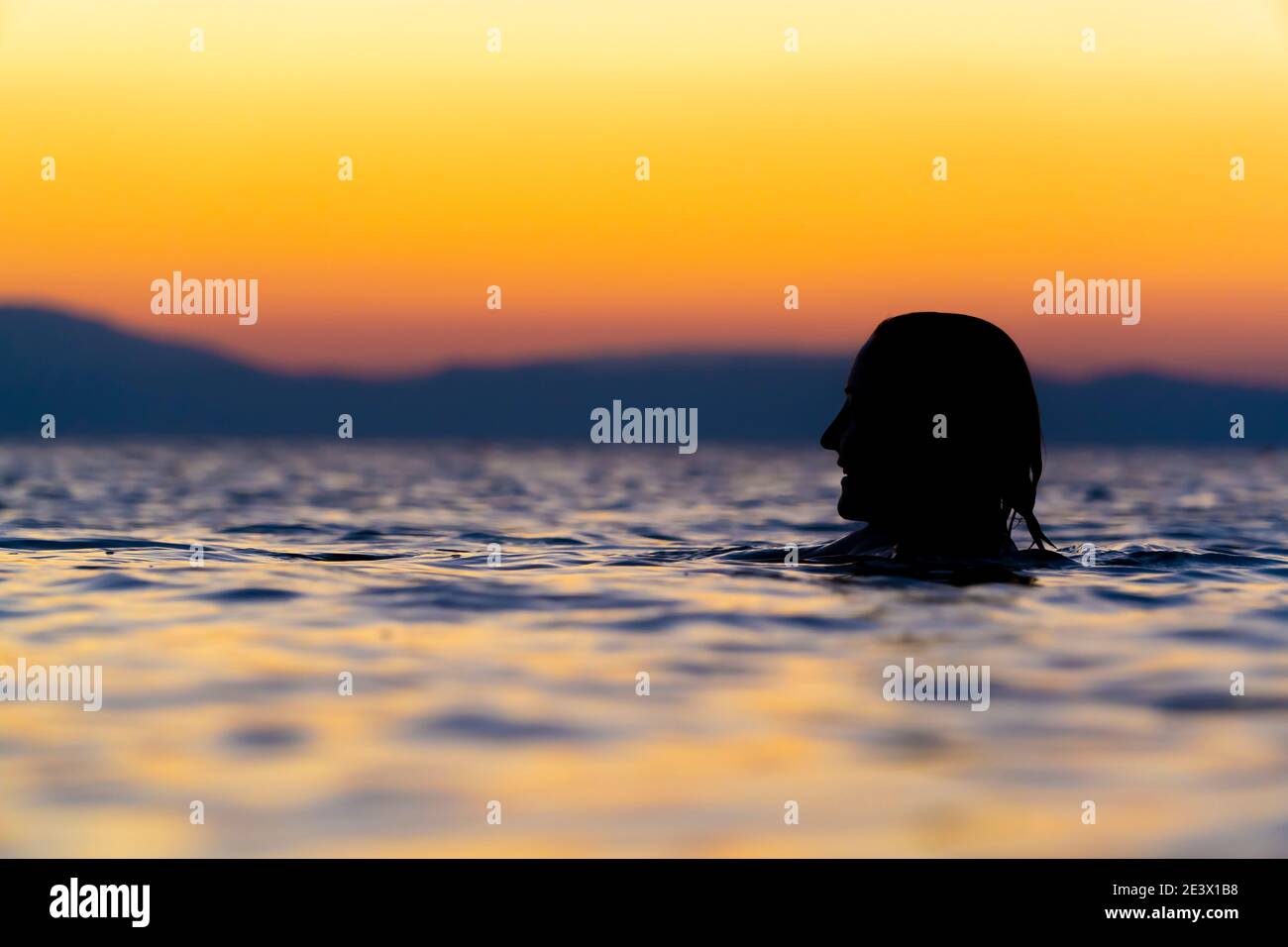 Fokussiere dich auf eine schwarze weibliche Kopfsilhouette, die bei Sonnenaufgang im Meer schwimmt. Die Frau lächelt. Orangefarbener Himmel und blaues Wasser mit flachem Fokus Stockfoto