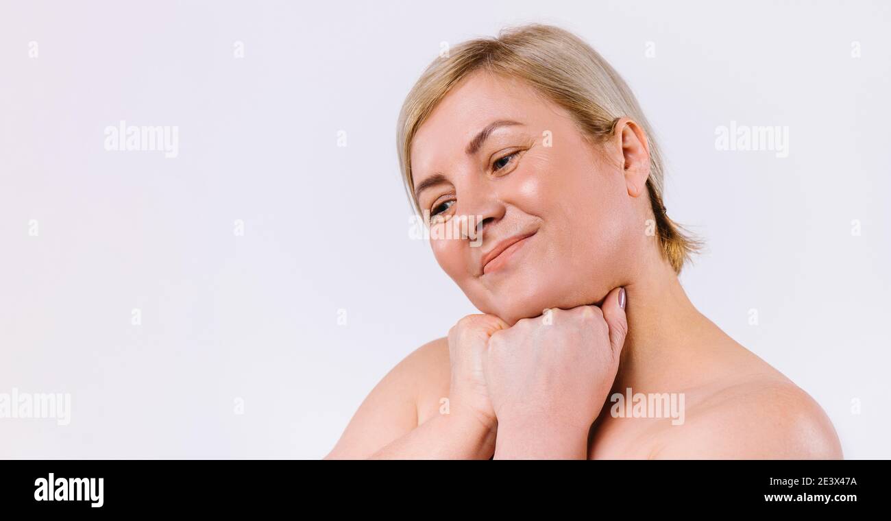 Banner. Eine nette ältere Frau mit sauberer und natürlicher Haut hält beide Hände unter das Gesicht und schaut nach unten. Skinimalismus auf weißem Hintergrund. Stockfoto