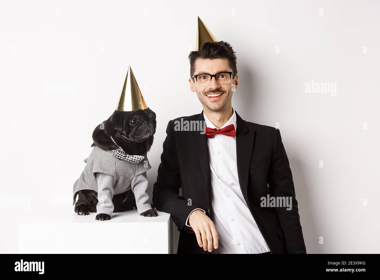 Bild von schönen jungen Mann Geburtstag feiern mit niedlichen schwarzen Mops in Party-Kostüm und Kegel auf dem Kopf, stehen über weißem Hintergrund Stockfoto