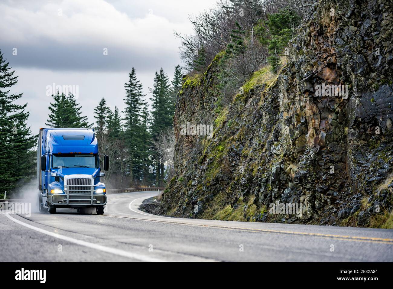 Big Rig leistungsstarke blaue Semi-Truck mit Rohren Grillschutz Transport von gewerblicher Fracht in Sattelaufliegern, die auf der Wicklung laufen Nasse Straße mit Regenstaub w Stockfoto