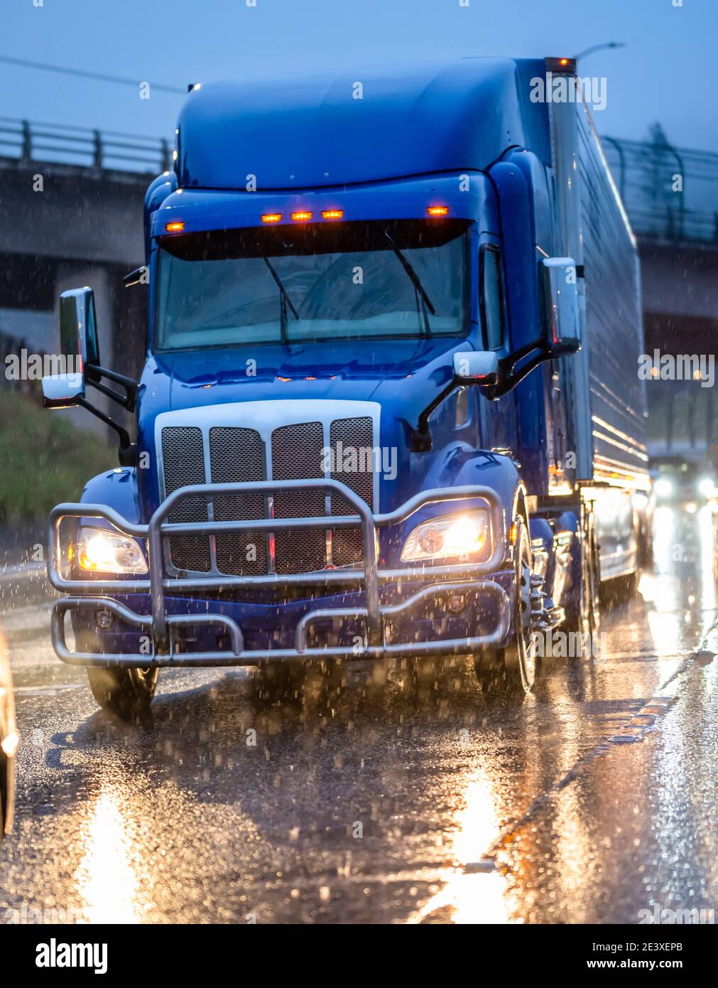 Big Rig stilvolle Industrie blau Semi-Truck mit eingeschaltet Scheinwerfer, die Ladung in Kühlraum-Sattelauflieger transportieren, die auf dem laufen Dämmerung nasse Straße Stockfoto