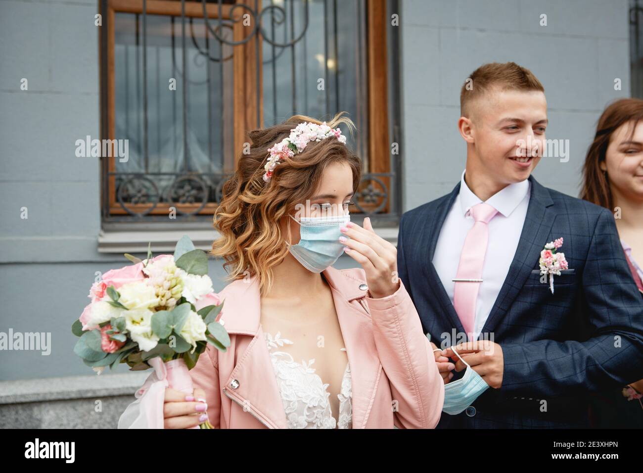 Die Braut in einer medizinischen Maske mit dem Bräutigam in der Nähe des Standesamtes. Hochzeit während der covid-19 Epidemie, Schutz vor dem Virus in öffentlichen Räumen. Stockfoto