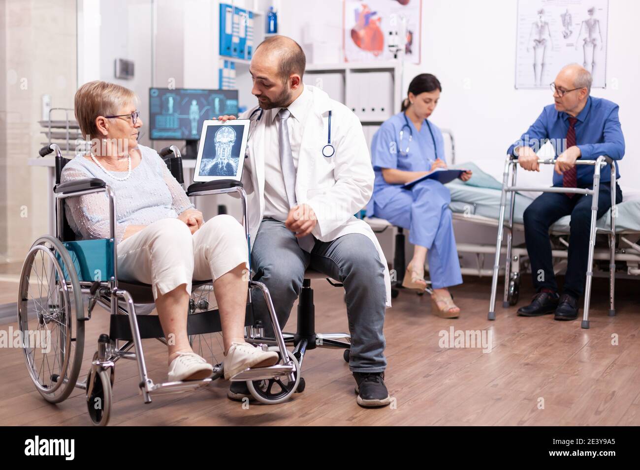 Behinderte ältere Frau mit Gehbehinderten sitzt im Rollstuhl und spricht mit dem Arzt über einen Tablet-pc. Mann mit Behinderungen, Gehrahmen sitzt im Krankenhausbett. Gesundheitssystem, Klinikpatienten. Stockfoto