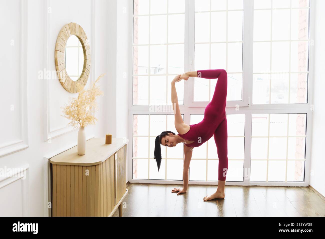 Porträt einer schönen jungen Frau in rosa Sportkleidung Yoga zu Hause in der Nähe des Fensters Durchführung einer Variation der Mondsichel Pose, Ardha Stockfoto