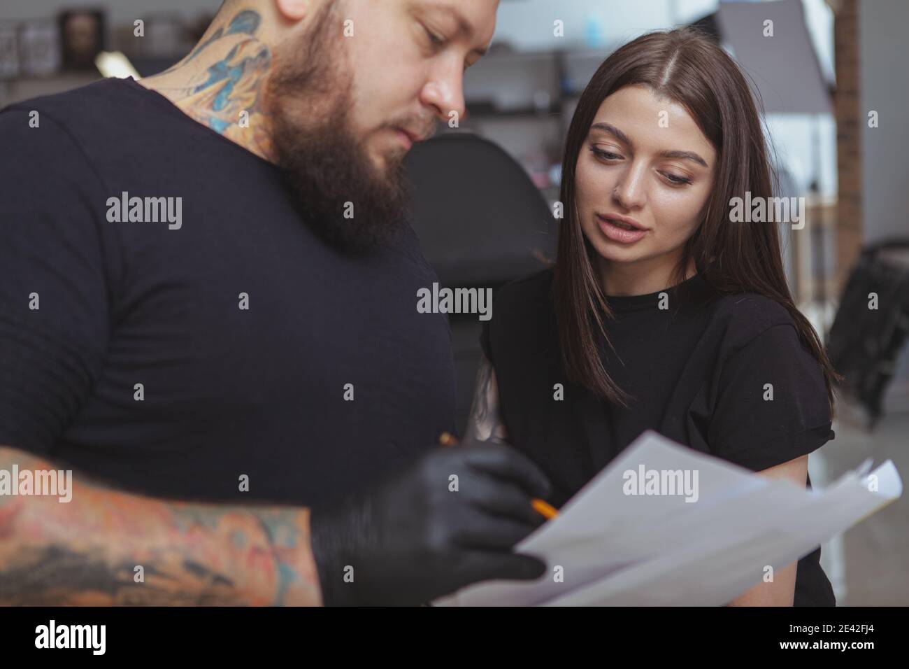 Zugeschnittenes Bild eines bärtigen professionellen Tattoo-Künstlers, der seiner Kundin dabei hilft, zu entscheiden, welches Tattoo sie erhalten soll. Charmante Frau Wahl neue Tätowierung bei der Stockfoto