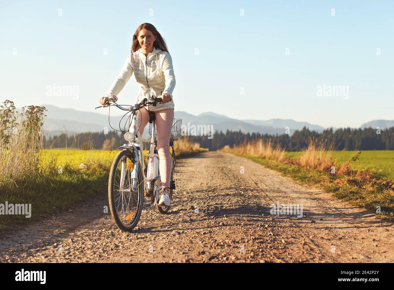 Junge Frau fährt Fahrrad auf staubigen Straße in Richtung Kamera, Nachmittag Sonne scheint auf Felder und Wald im Hintergrund Stockfoto