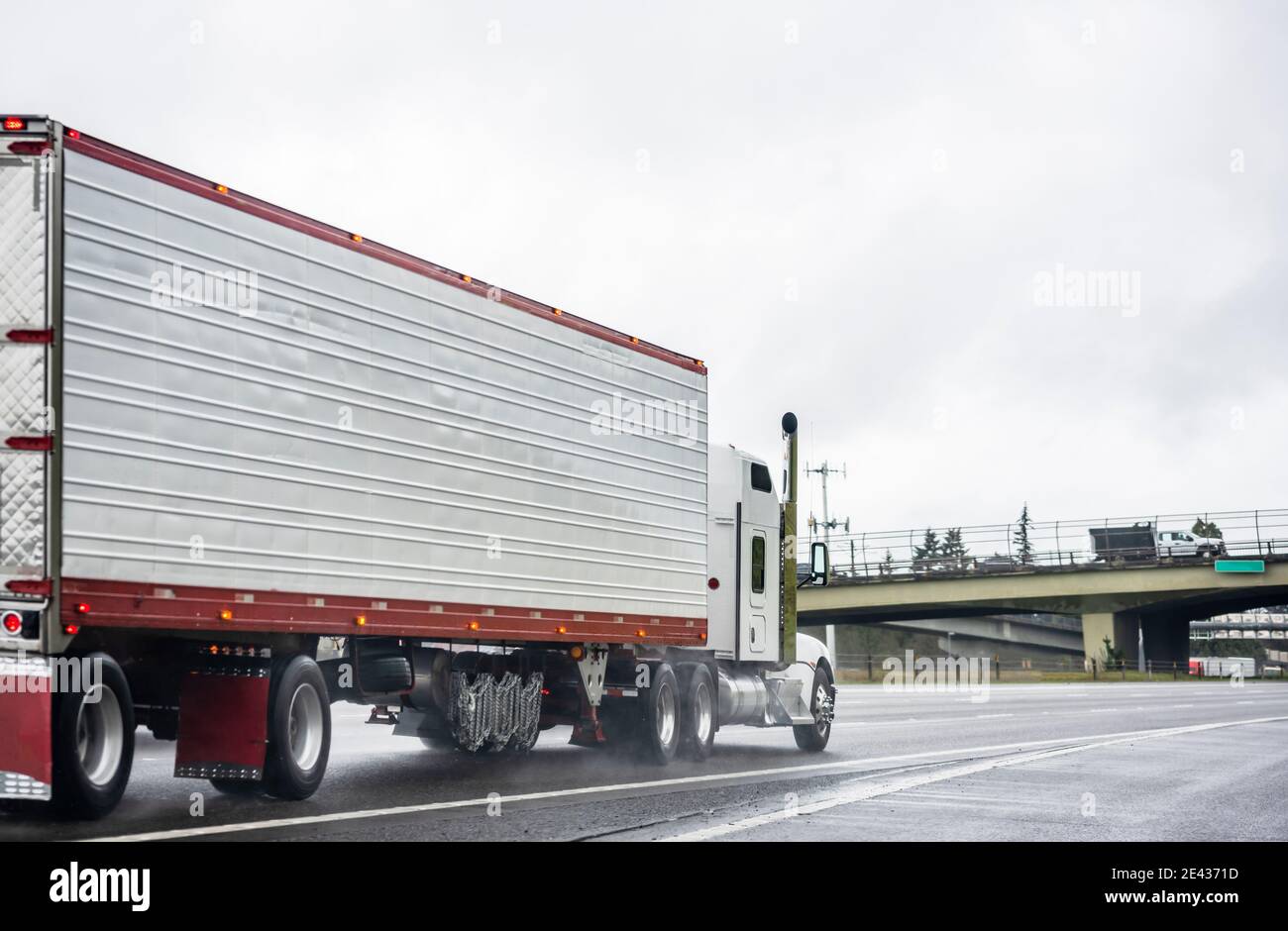Big Rig stilvolle Industrie weißen Semi-Truck mit eingeschaltet Scheinwerfer, die Ladung in Kühlraum-Sattelauflieger transportieren, die auf dem laufen Dämmerung nasse Straße Stockfoto