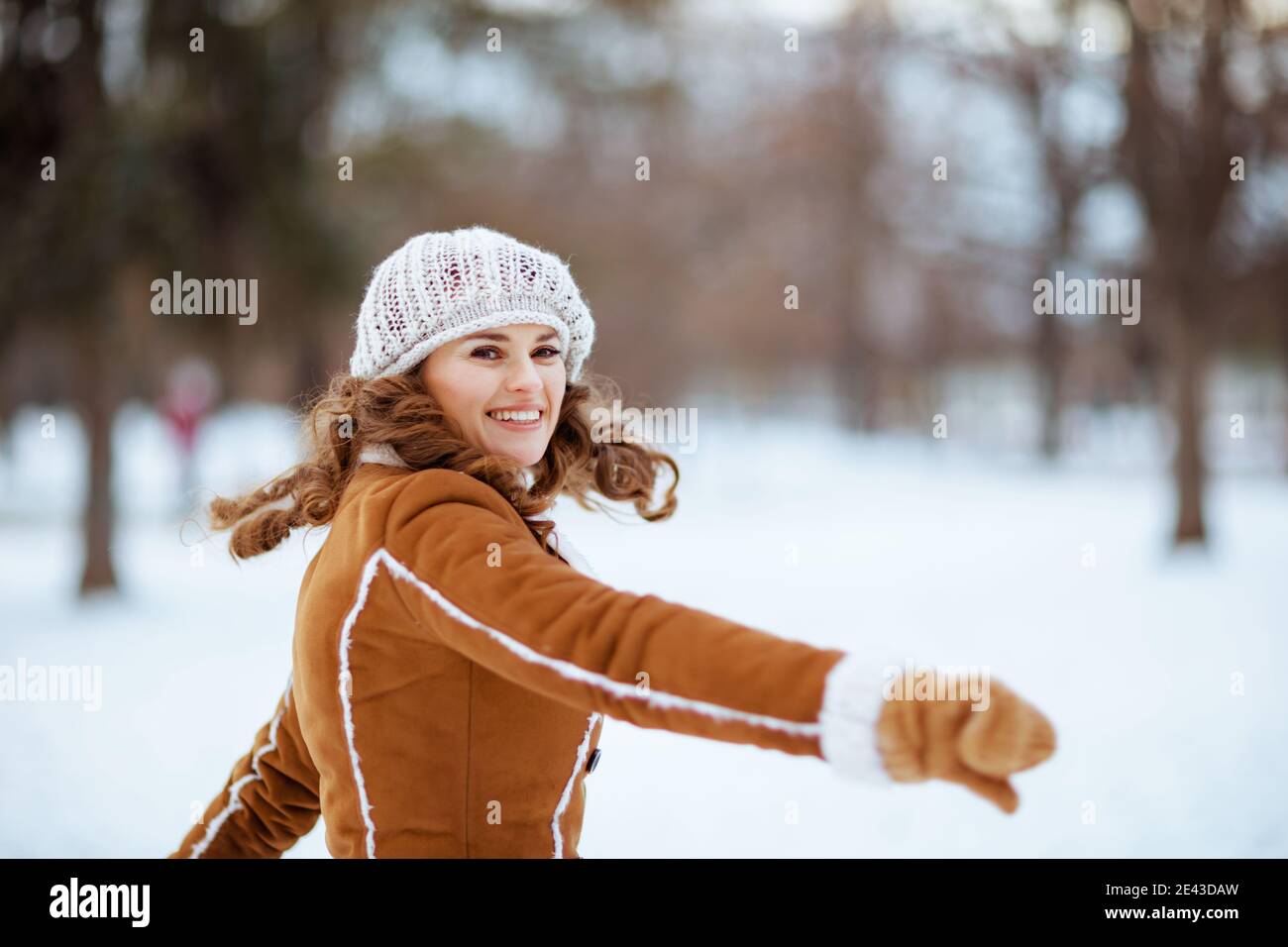 Lächelnd elegante Frau mittleren Alters mit Fäustlingen in einem gestrickten Hut und Schaffell Mantel jubeln im Freien im Stadtpark im Winter. Stockfoto