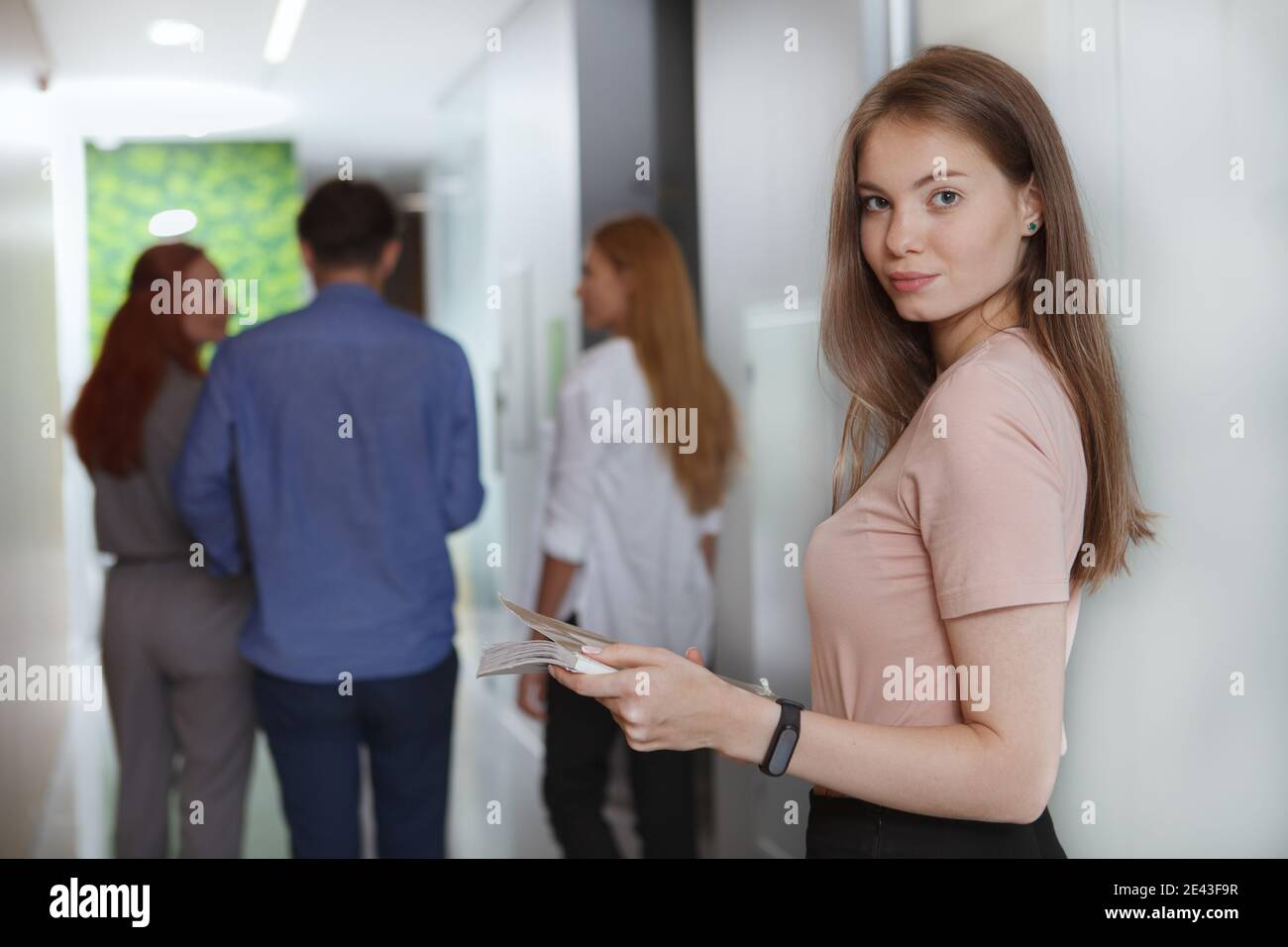 Attraktive junge Geschäftsfrau lächelt zur Kamera, ihre Kollegen gehen im Hintergrund durch den Flur Stockfoto