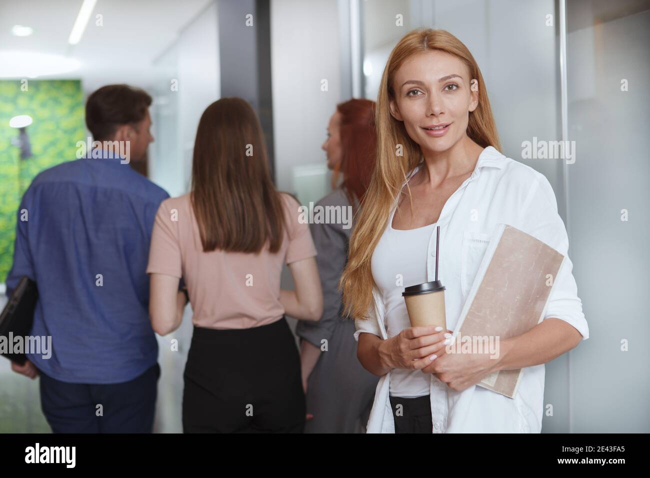 Selbstbewusste schöne Geschäftsfrau lächelt an der Kamera, Kaffee bei der Arbeit, ihre Kollegen gehen auf Hintergrund Stockfoto