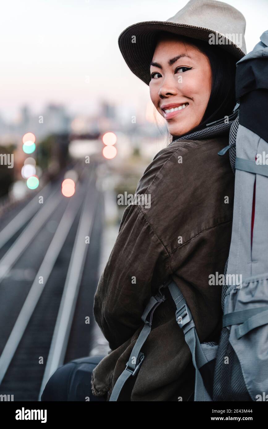 Rückansicht der erfreut asiatische weibliche Backpacker sitzt auf Hintergrund Von Eisenbahn und Blick auf Kamera über Schulter während des Abenteuers In Australien Stockfoto