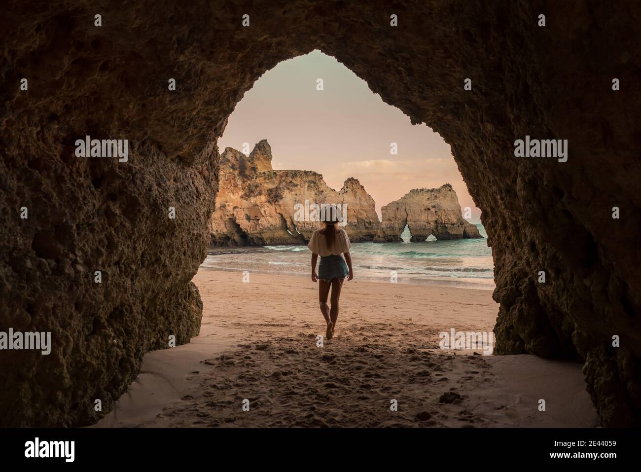 Rückansicht einer nicht erkennbaren Frau in legerer Kleidung und Hut auf Sand im Eingang der Höhle in der Nähe des Meeres bei Sonnenuntergang in Benagil Höhlen in Algarve, Port Stockfoto