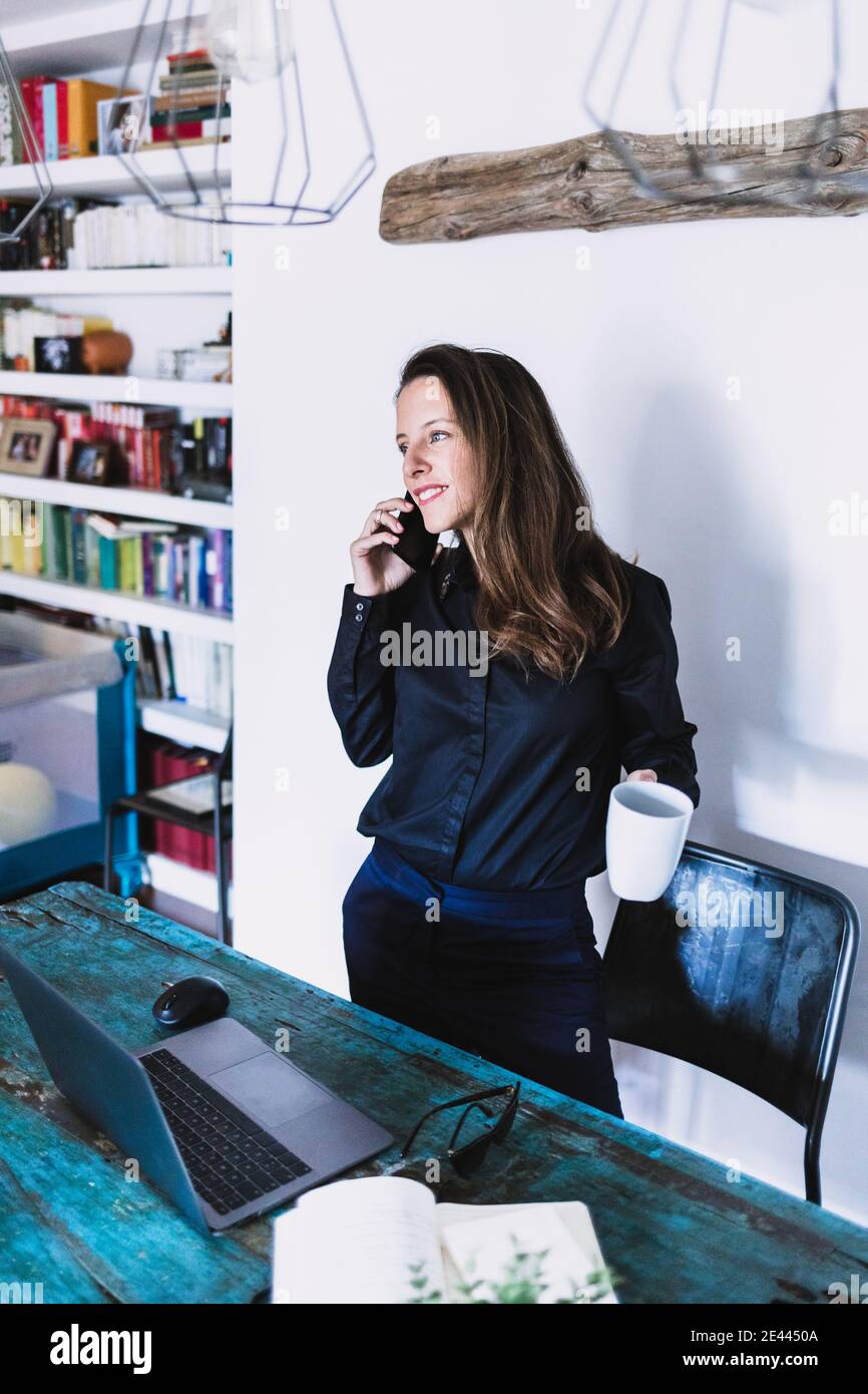Lächelnd junge Frau mit einer Tasse Kaffee in der Hand antworten Telefonanruf, während Sie in der Nähe des Tisches mit Laptop während der Fernbedienung stehen Arbeiten Sie von zu Hause aus Stockfoto