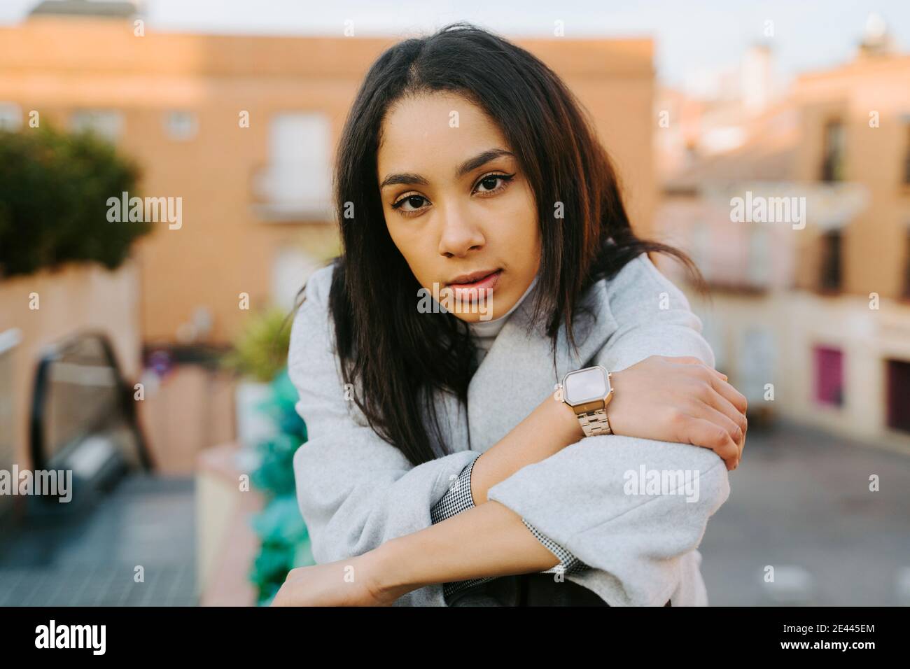 Selbstbewusst junge Frau trägt modischen blauen Mantel und Blick auf Kamera sitzt auf Steinzaun in der Stadt Stockfoto