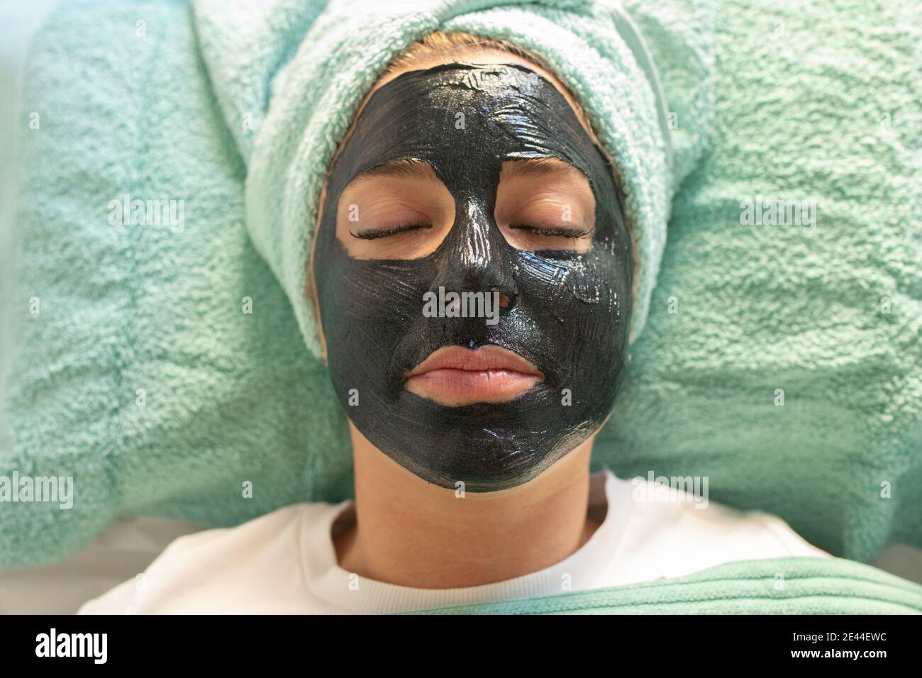 Nicht erkennbare junge weibliche Klientin in legerer Kleidung mit Handtuch auf Kopf und schwarze Tonmaske im Gesicht Stockfoto