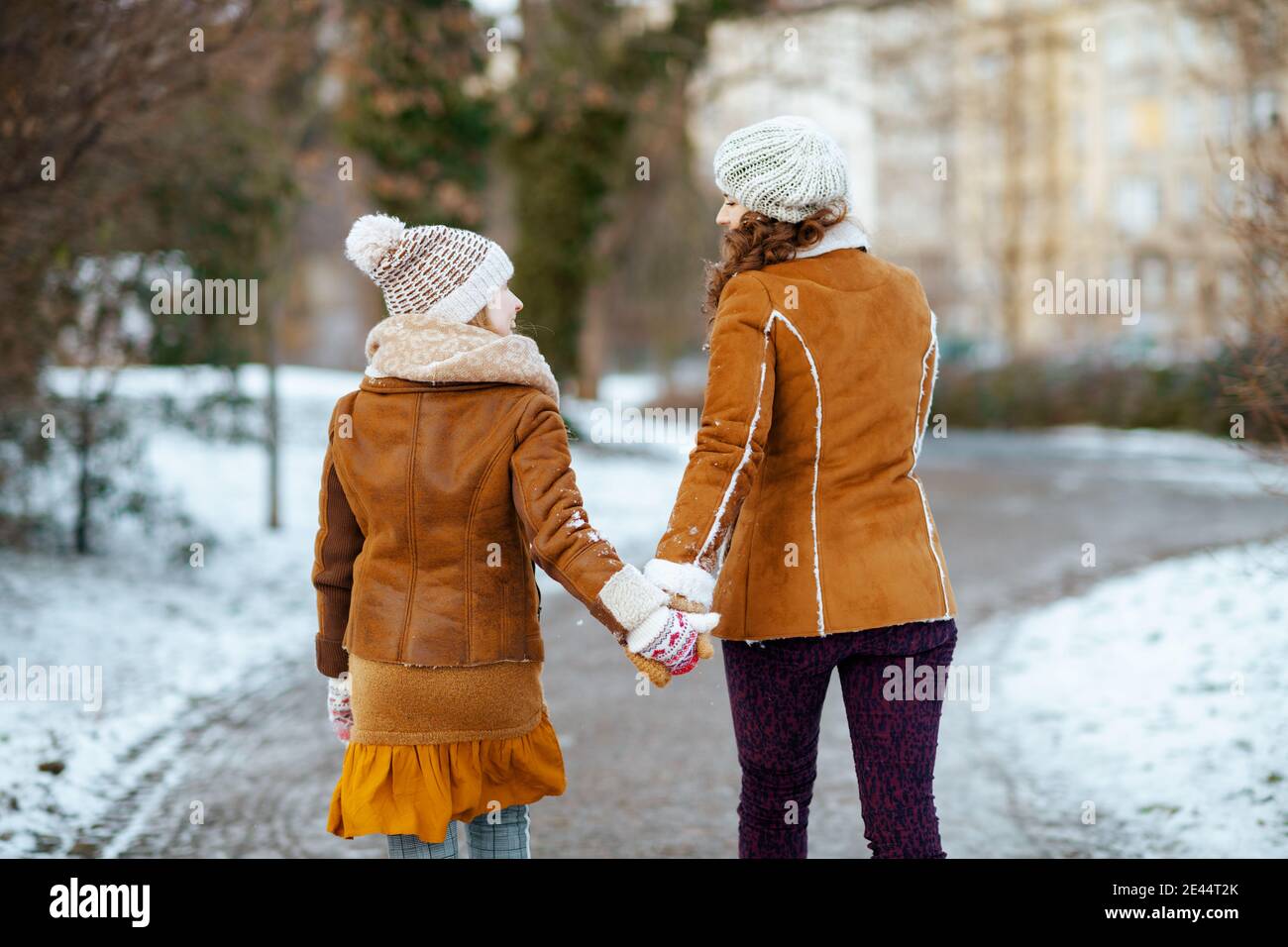 Von hinten gesehen Mutter und Kind in einem gestrickten Hüte Und Schaffell Mäntel mit Fäustlingen in einem gestrickten Hut und Schaffell Mantel zu Fuß im Freien in der Stadt Stockfoto