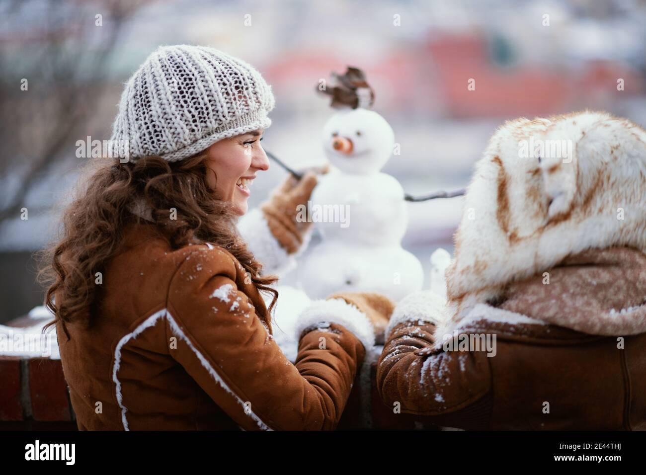 Von hinten gesehen Mutter und Tochter in einem gestrickten Hüte und Schaffell Mäntel draußen im Stadtpark im Winter machen einen Schneemann. Stockfoto