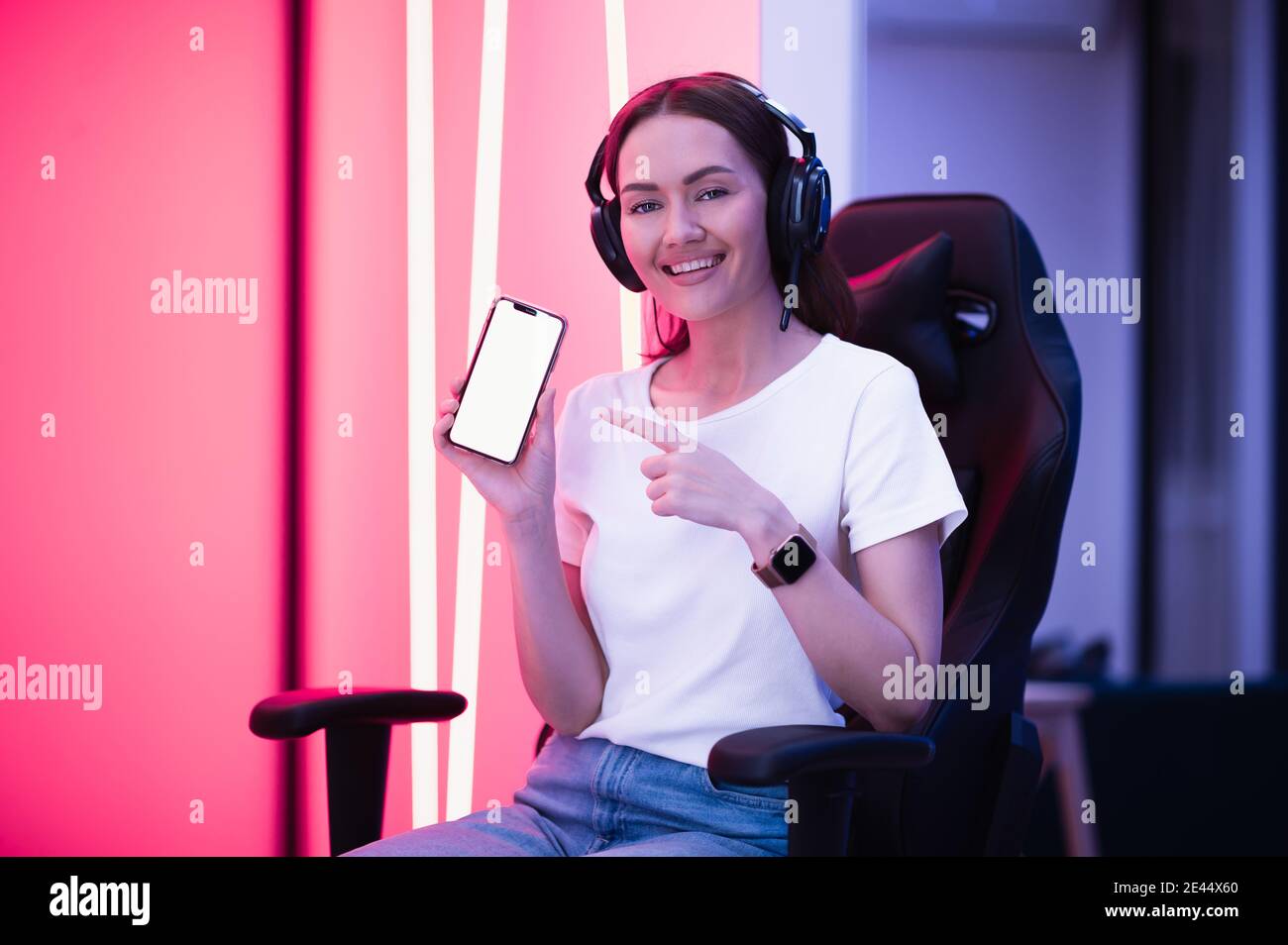 Lächelndes Gamer Mädchen trägt Headset zeigt Smartphone und zeigt auf leeren Bildschirm mit ihrem Finger. Stockfoto