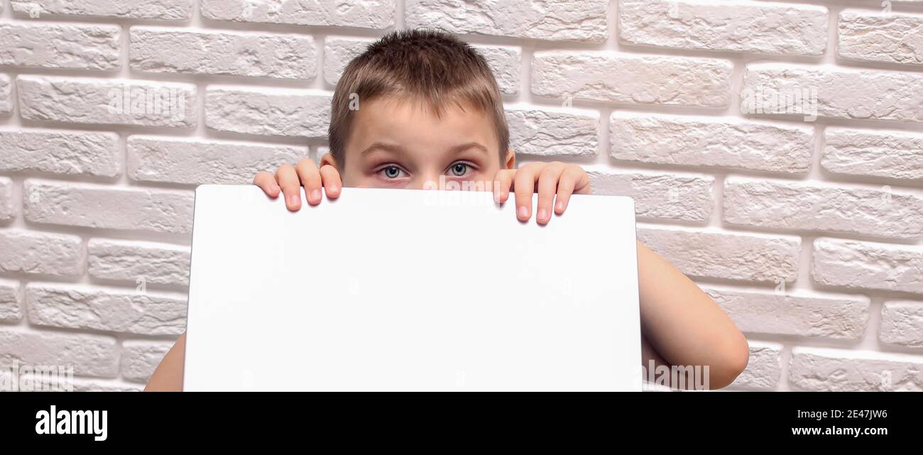 Der Junge schaut hinter seinem Laptop heraus. Auf dem Hintergrund einer weißen Backsteinmauer. Stockfoto