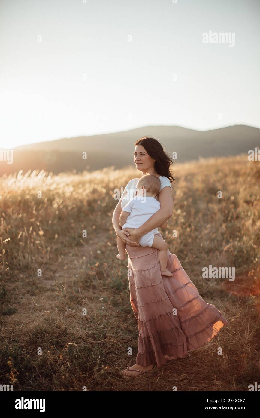 Mutter hält ihr gebreches Baby auf Feldern und schaut in den Sonnenuntergang Stockfoto