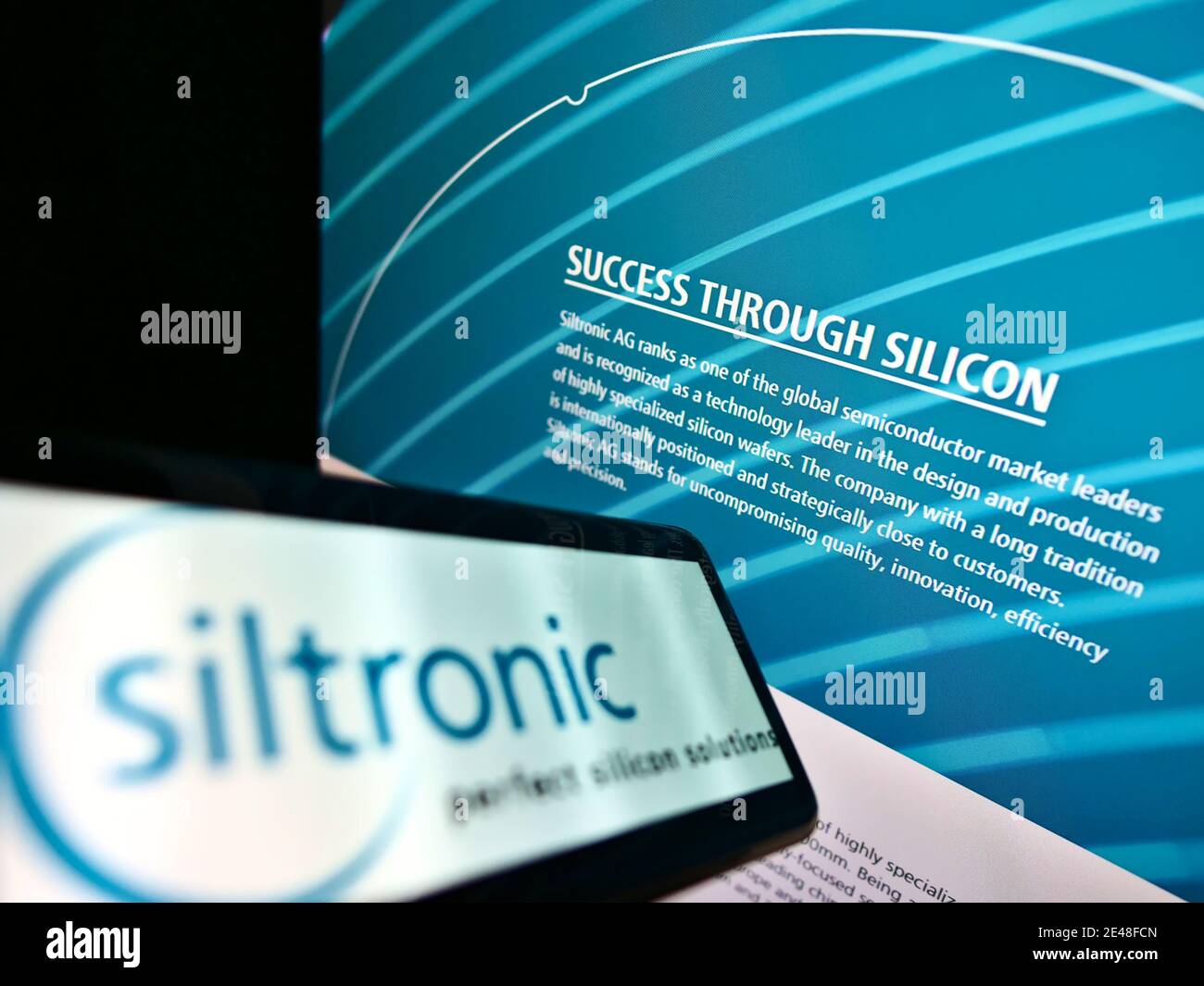 Mobiltelefon mit Logo des deutschen Siliziumwaferherstellers Siltronic AG auf dem Bildschirm vor der Website. Focus on Slogan auf Monitor im Hintergrund. Stockfoto