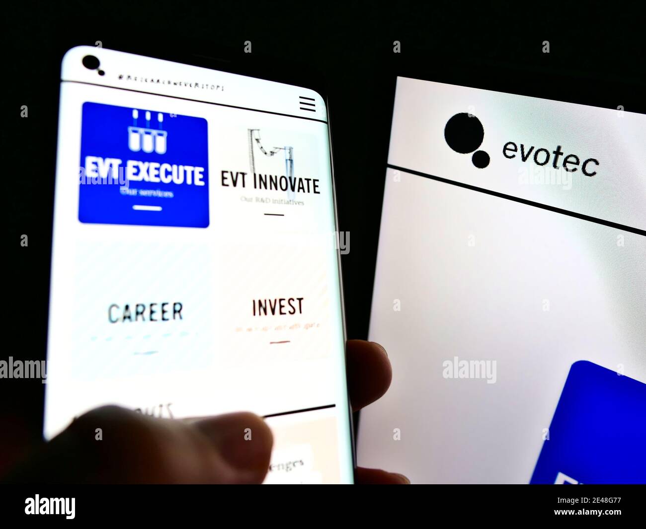 Person, die Smartphone mit Website des deutschen Biotechnologie-Unternehmens Evotec SE neben Computer-Monitor hält. Konzentrieren Sie sich auf das Unternehmenslogo auf dem rechten Bildschirm. Stockfoto