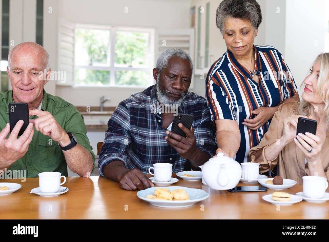 Ältere kaukasische und afroamerikanische Paare sitzen am Tisch und trinken Tee mit Smartphone zu Hause Stockfoto