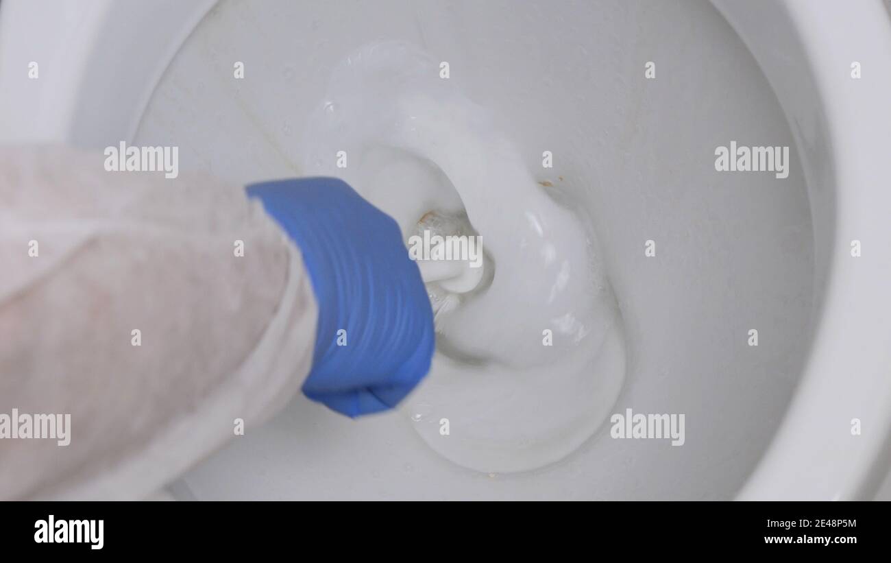 Eine Person mit Schutzkleidung und Handschuhen, die Toilette im Badezimmer mit einer Toilettenbürste desinfizieren Stockfoto