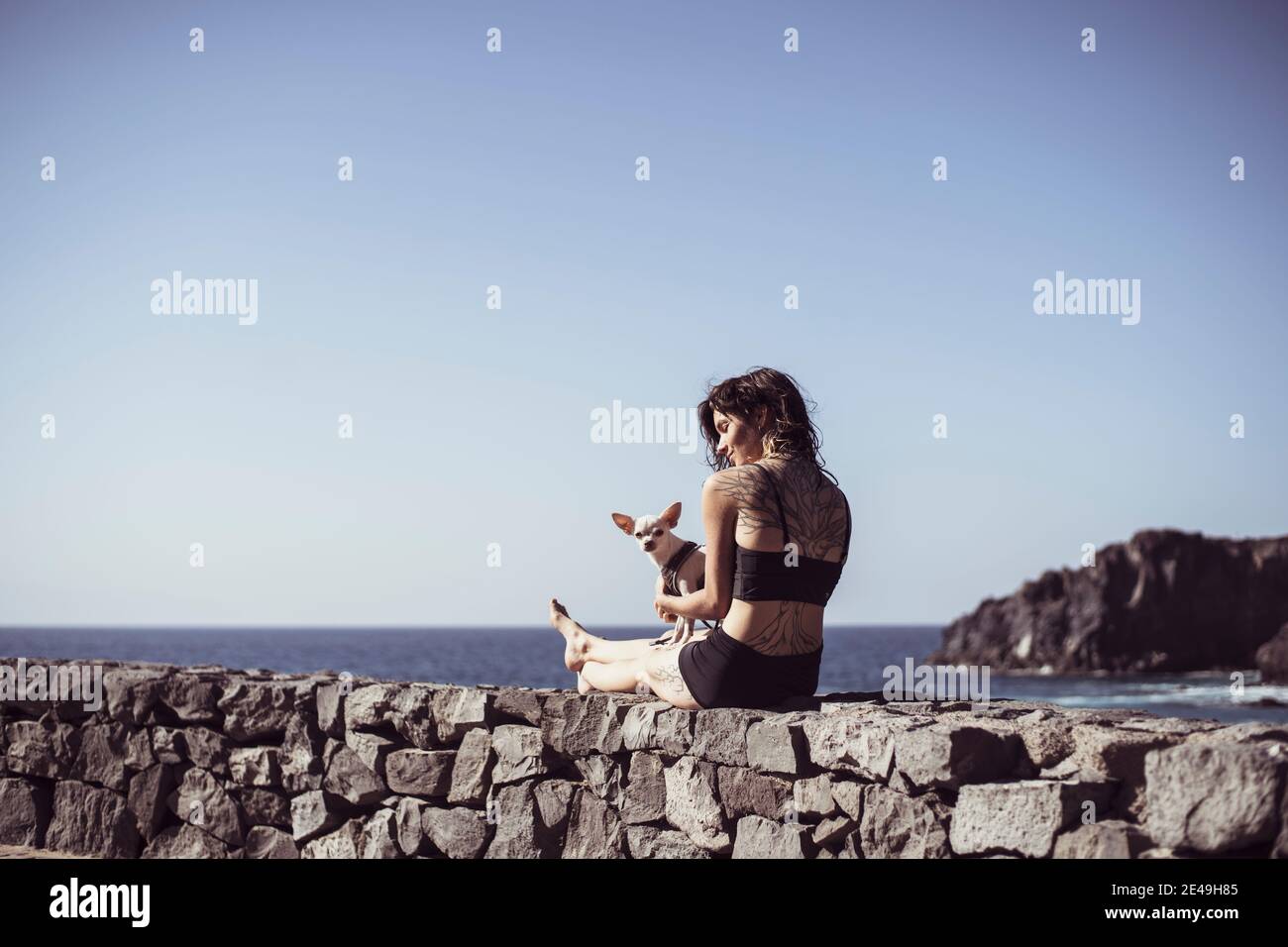 Gesund fit Frau mit Tattoos sitzt am Meer mit in Sonne mit kleinem Hund Stockfoto