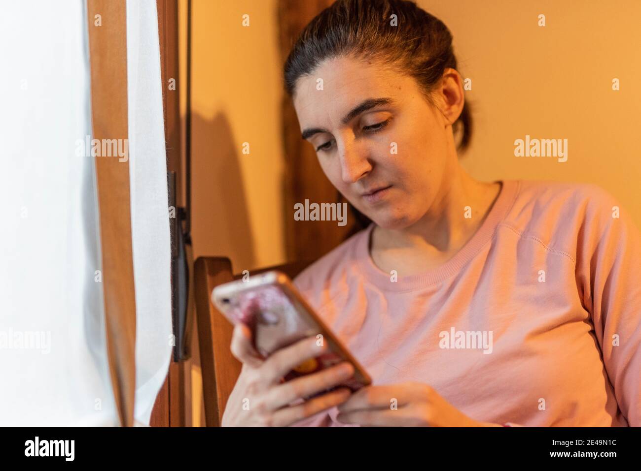 Frau mit Handy neben einem Fenster mit natürlichen Helles und warmes Licht Stockfoto