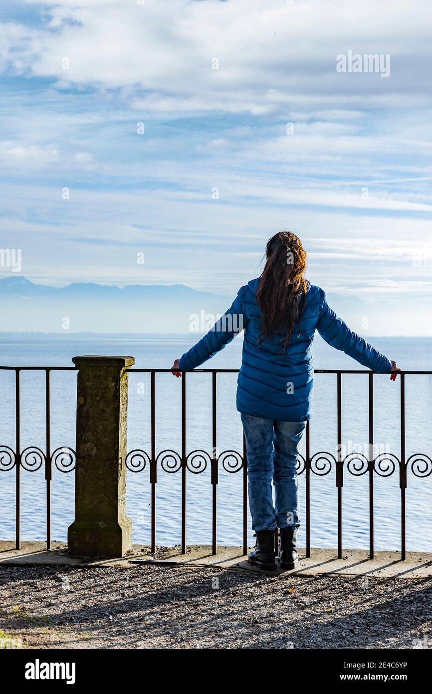 Frau in einer blauen Daunenjacke auf dem Geländer vorbei Ein See Stockfoto