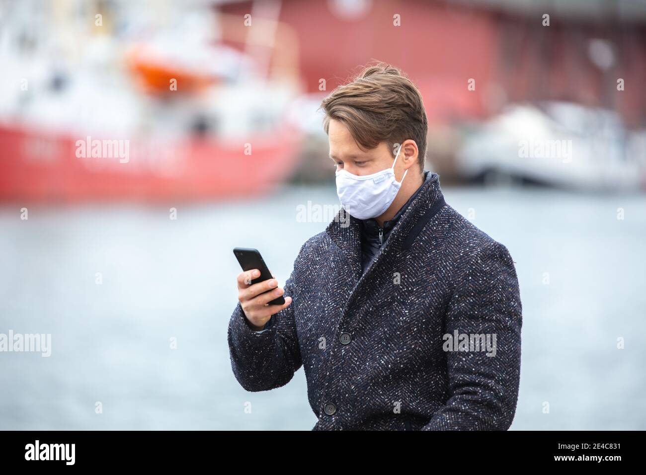 Junger Mann Corona Zeit, mit Alltagsmasken, in der kalten Jahreszeit in der Stadt. Mit seiner Mund- und Nasenmaske an öffentlichen Plätzen. Schaut auf sein Smartphone. Stockfoto