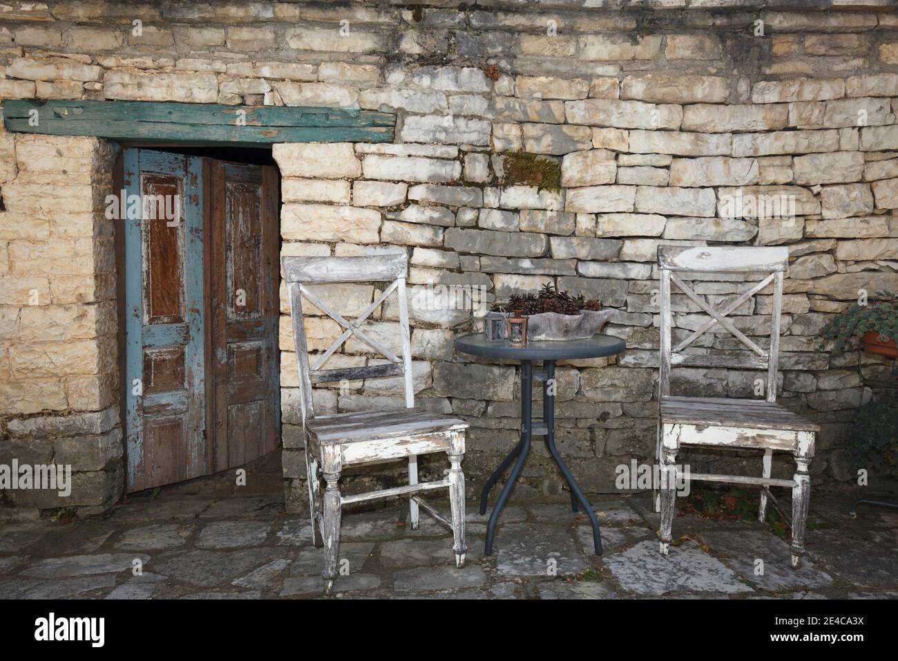 Milieu mit Tisch, zwei Holzstühlen, verwitterter Haustür und rauem Mauerwerk in Monodendri, Zagori-Dörfern, Pindus-Bergen, Epirus, Nordgriechenland Stockfoto