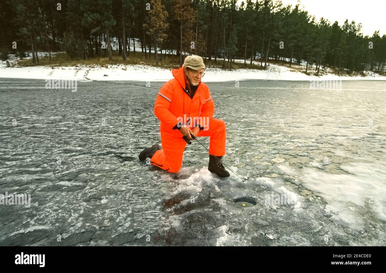 Ein begeisterter Winterfischer beobachtet seine Angelschnur mit einem gelben Bobber, der in einem Loch im Eis auf dem gefrorenen Dickey Lake im Kootenai National Forest schwimmt, einer riesigen Wildnis, die das ganze Jahr über für Outdoor-Erholung im Nordwesten von Montana, USA, beliebt ist. Dieser erfahrene Angler mit kaltem Wetter trägt warme Kleidung unter farbenfroher, wasserdichter Oberbekleidung und Stiefel mit Stollen zum Wandern auf der rutschigen Eisfläche des Sees. Stockfoto