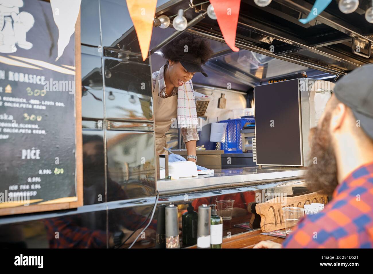 Junge afro-amerikanische Mitarbeiterin in Fast-Food-Service schreiben Der Auftrag von einem jungen kaukasischen bärtigen Hipster mit einem Kap Stockfoto
