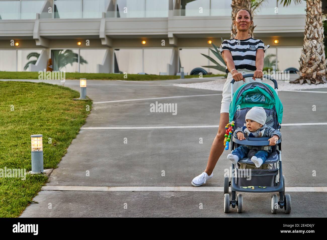 Ganzkörper froh Frau mit niedlichen Baby im Kinderwagen stehen Auf dem Weg vor dem modernen Hotel am Sommerabend im Resort Stockfoto