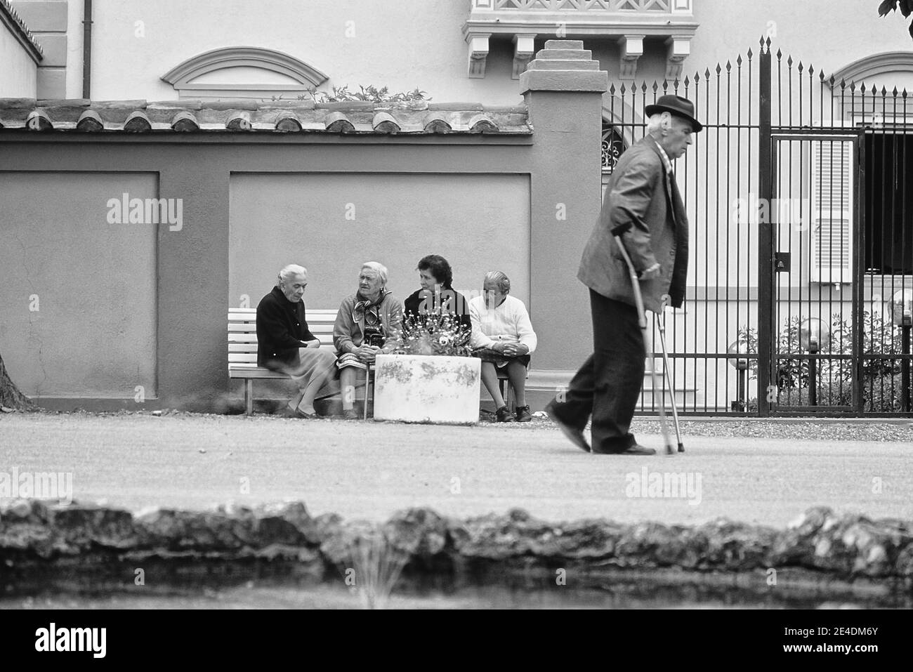 Ein älterer Mann mit Krücken, der an vier älteren italienischen Damen vorbeifährt, die auf einer öffentlichen Bank sitzen und eine Diskussion führen. Italien Stockfoto