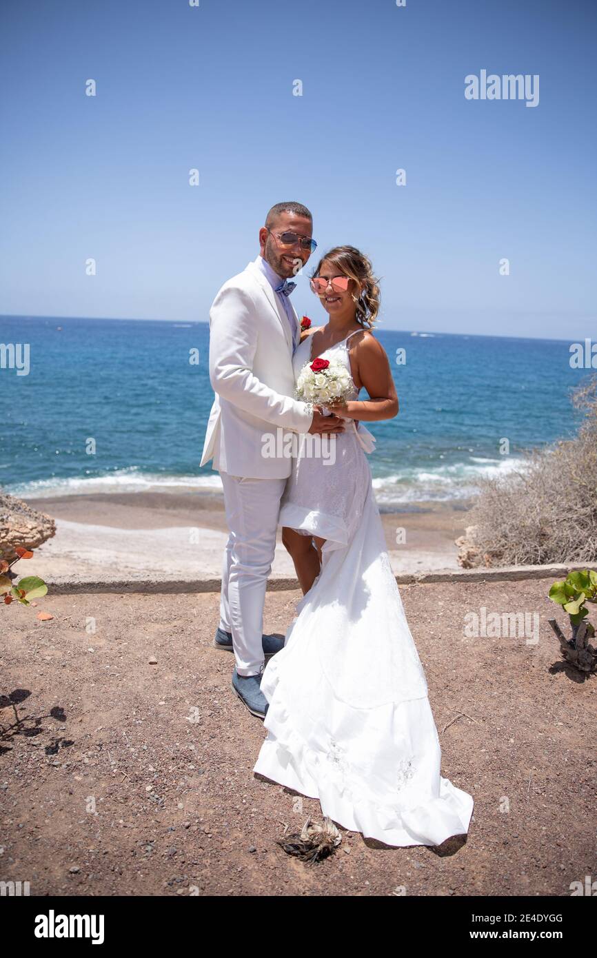 Das frisch verheiratete Paar hält den Brautstrauß hoch, sie sind elegant in Weiß gekleidet mit Sonnenbrillen und haben das Meer hinter sich. Pho. In voller Länge Stockfoto