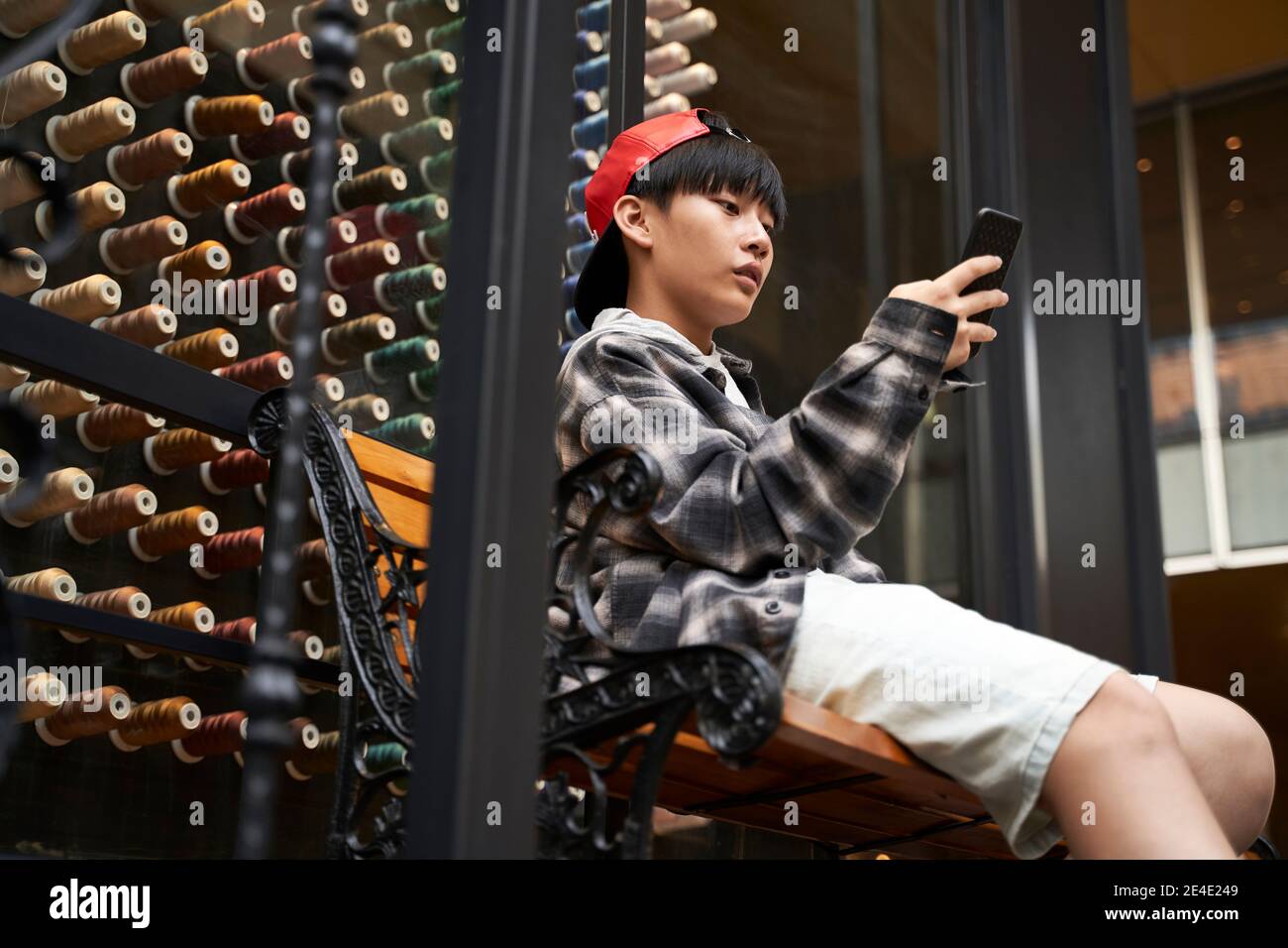 15-jähriger asiatischer Teenager-Junge, der auf der Bank sitzt und Handy anschaut Stockfoto