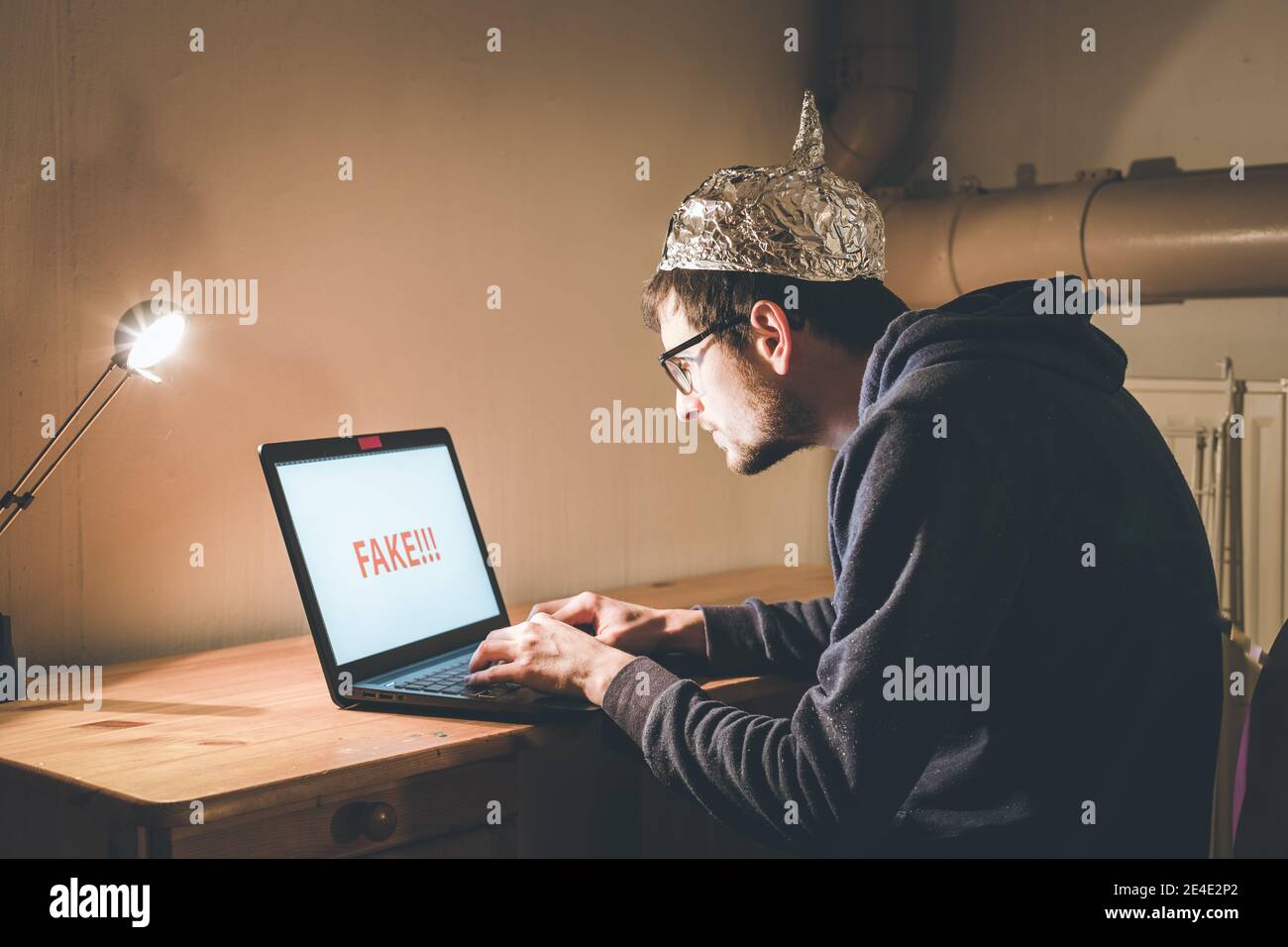 Der junge Mann mit Aluminiumkappe sitzt im dunklen Keller vor einem Laptop. Verschwörungstheorie Konzept Stockfoto