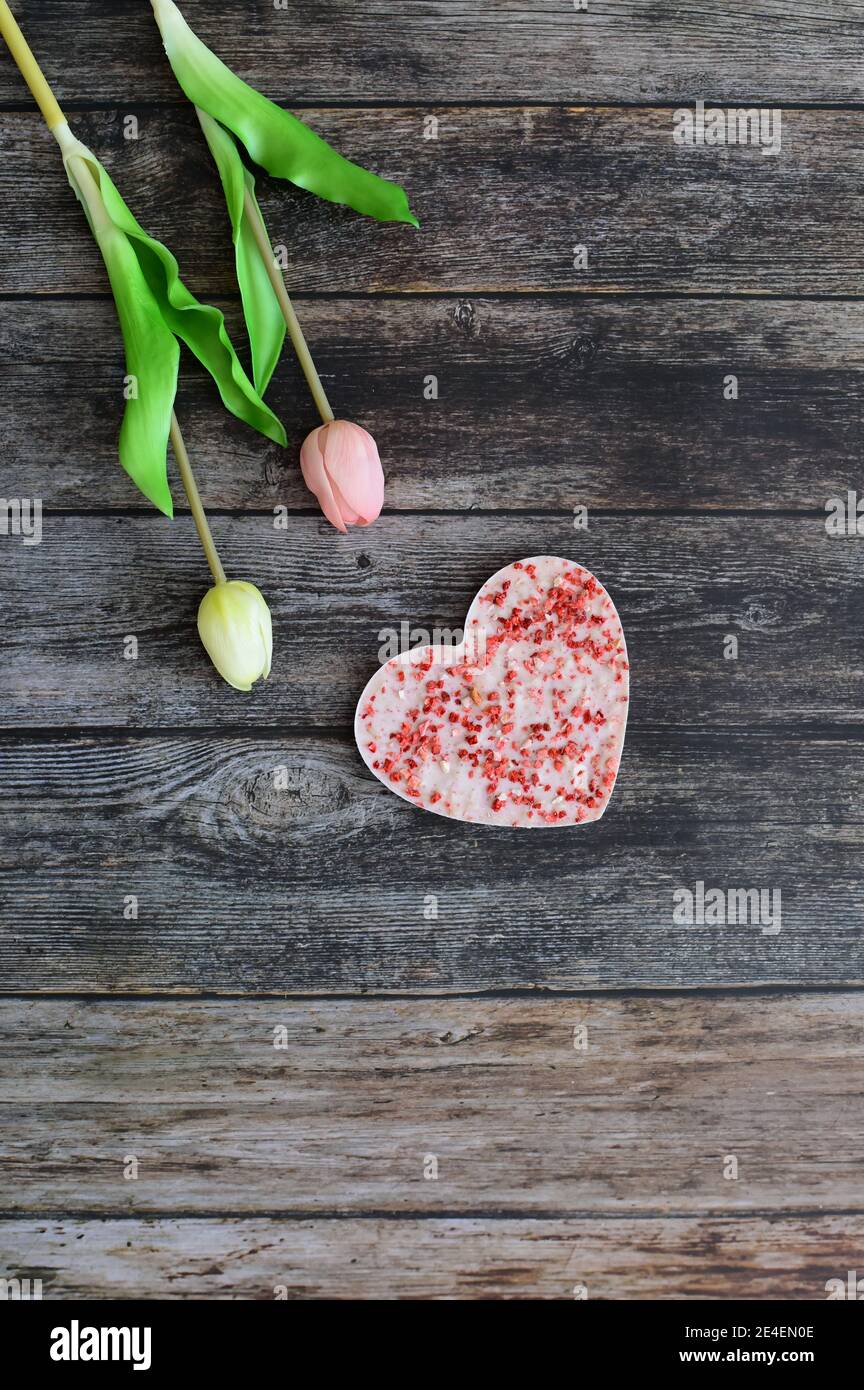 Eine herzförmige rosa Schokolade mit getrockneten Erdbeeren auf einem Vintage dunklen Holztisch Hintergrund mit Tulpenblüten. Liebe und Grußkarte Konzept. Stockfoto