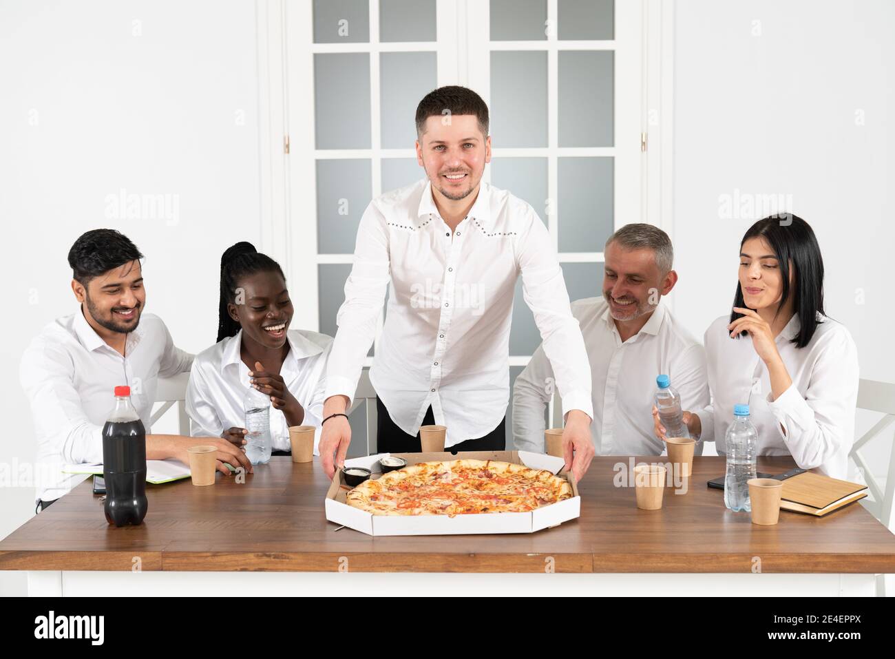 Das Business Team isst Pizza im Büro und sitzt um den Tisch. Überglücklich gemischte Mitarbeiter der Rennfirma versammelten sich am Bürotisch Stockfoto