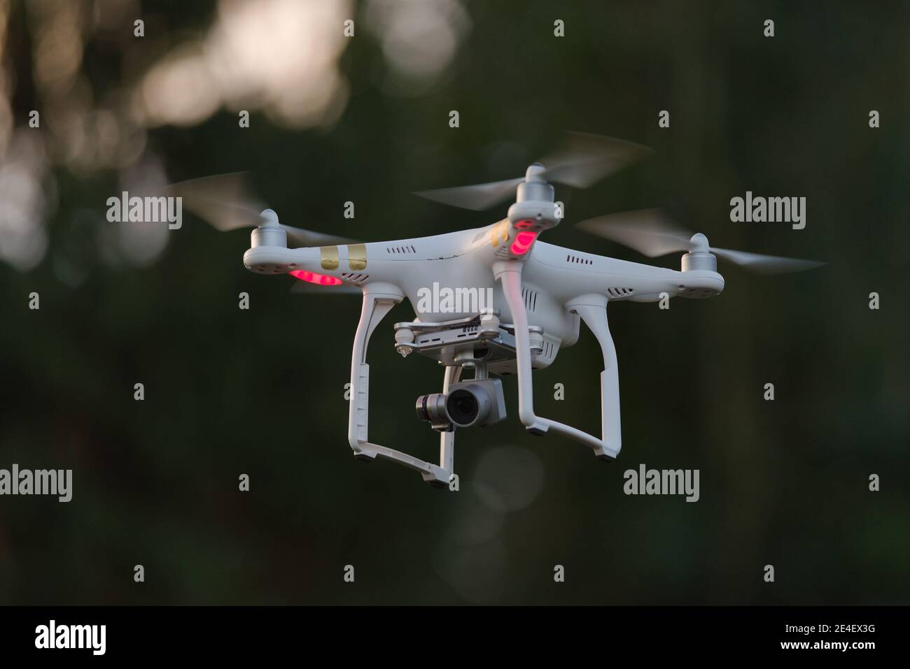 12 - Weiße Quadrotor-Drohne mit Orientierungslicht, die isoliert von dunklem unfokussiertem Hintergrund schwebt. Professionelle Kamera auf Gimbal unten gibt FPV Stockfoto