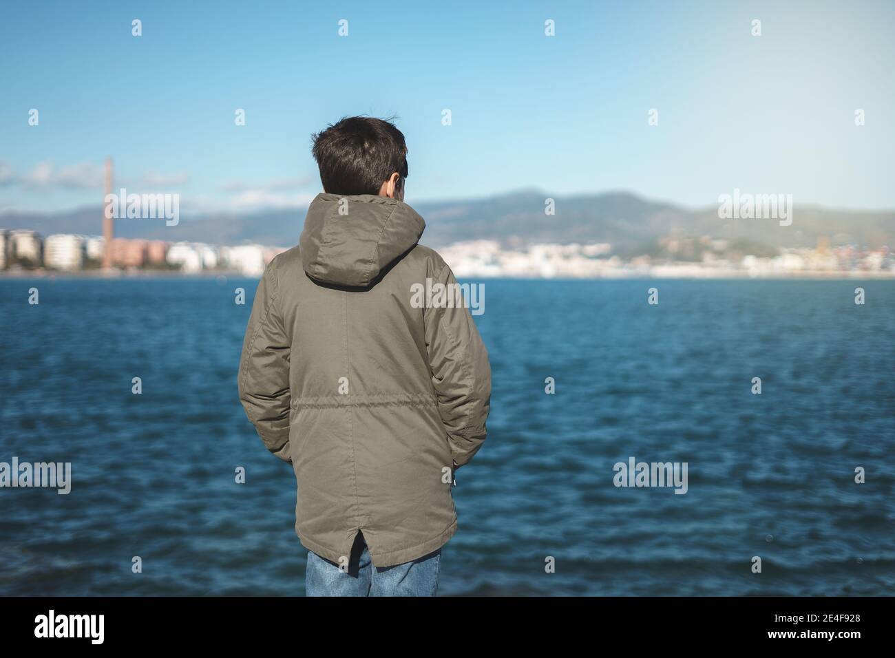 Teenager mit Blick auf das Meer in grünem Mantel, mit der Stadt im Hintergrund. Stockfoto