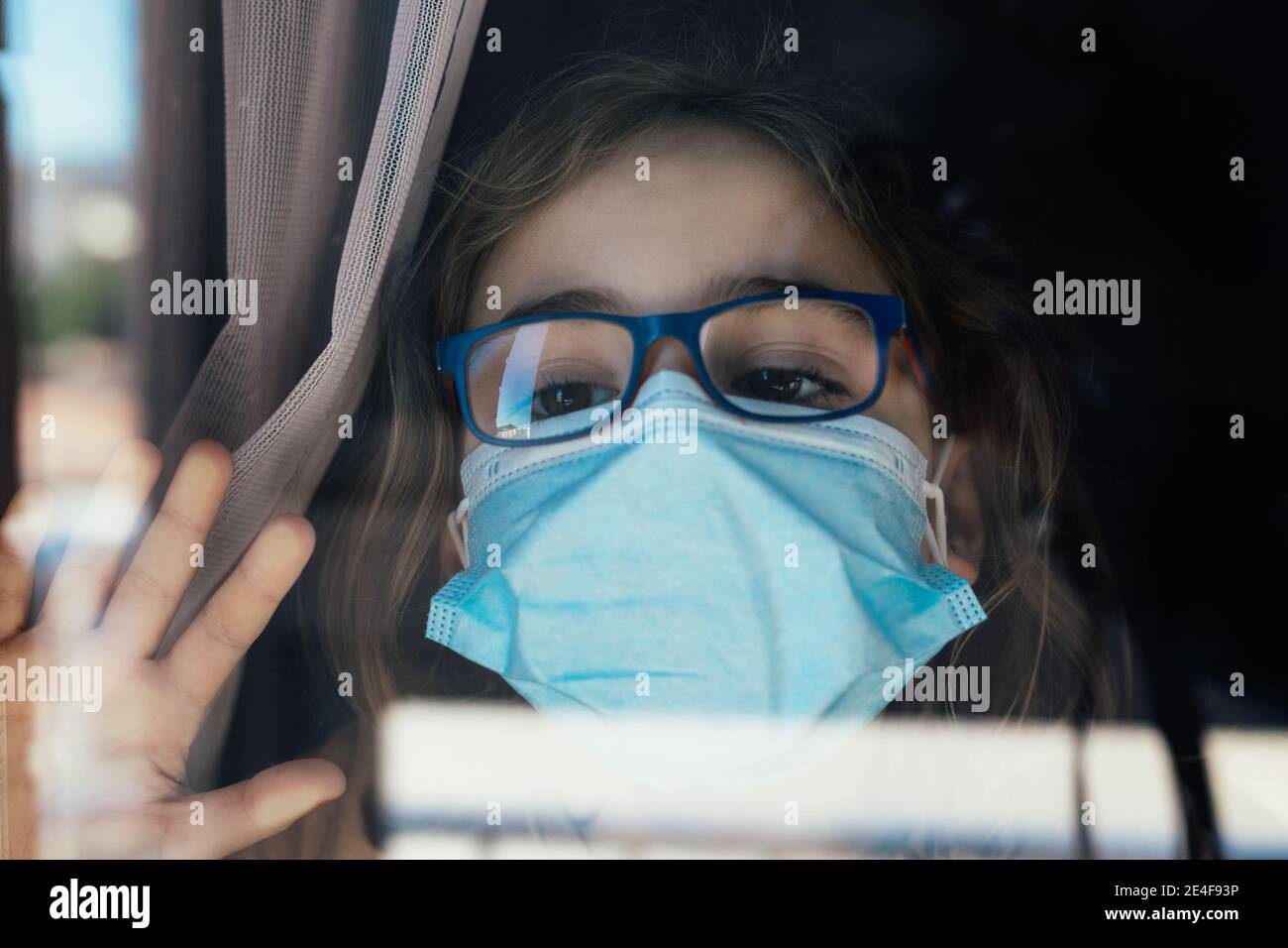 Mädchen mit Brille und Maske hinter einem Fenster, Hausbeengung, Coronavirus-Konzept Stockfoto