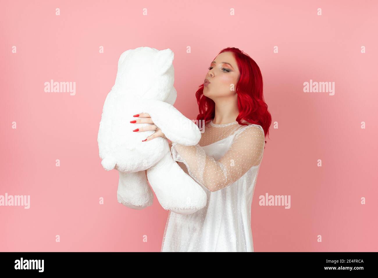 Close-up eine niedliche junge Frau in einem weißen Kleid und Mit roten Haaren bläst einen Kuss auf ein großes Weiß teddybär isoliert auf einem rosa Hintergrund Stockfoto