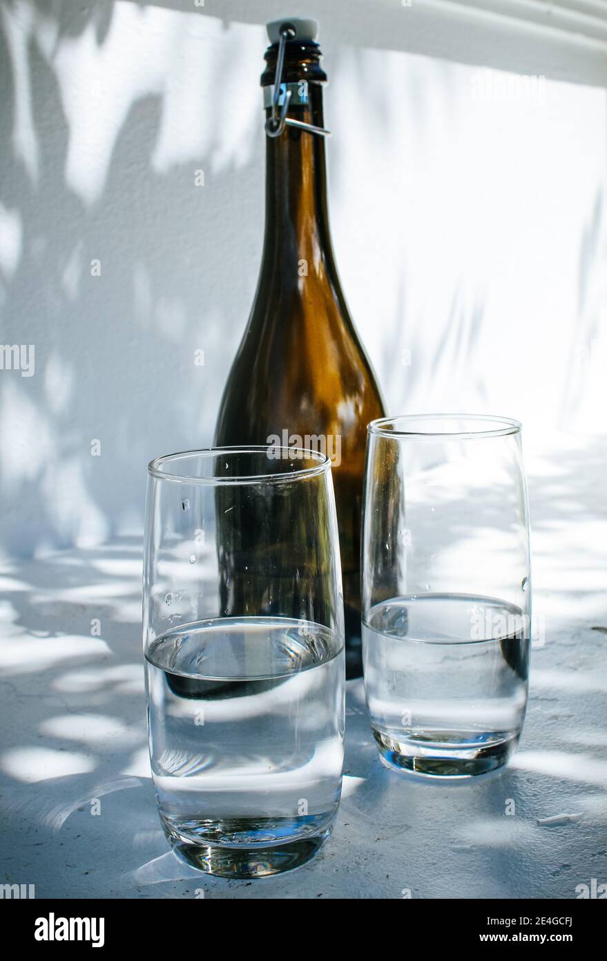 Eine Flasche und zwei Gläser Wasser stehen auf einem Weißer Rand gegen eine helle weiße Wand mit Schatten Blätter an einem sonnigen Sommertag Stockfoto