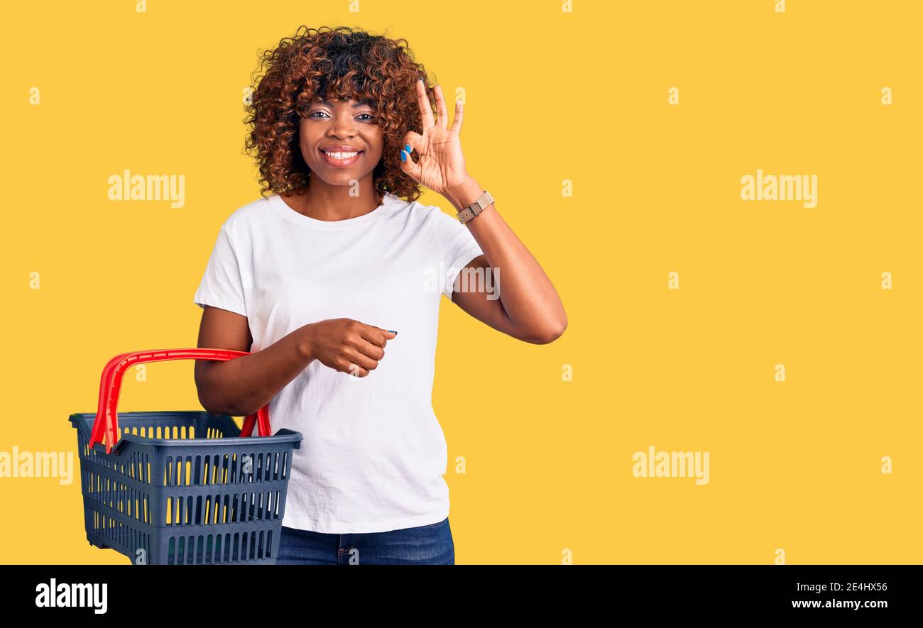 Junge afroamerikanische Frau hält Supermarkt Warenkorb tun ok Zeichen mit Fingern, lächelnd freundlich gestikulieren ausgezeichnete Symbol Stockfoto