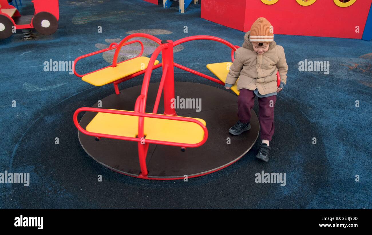 Liebenswert Kleinkind Junge in warmen Hut und Jacke reitet auf Kleine lustige Runde auf einem modernen Spielplatz im Garten des Apartmentgebäudes Stockfoto